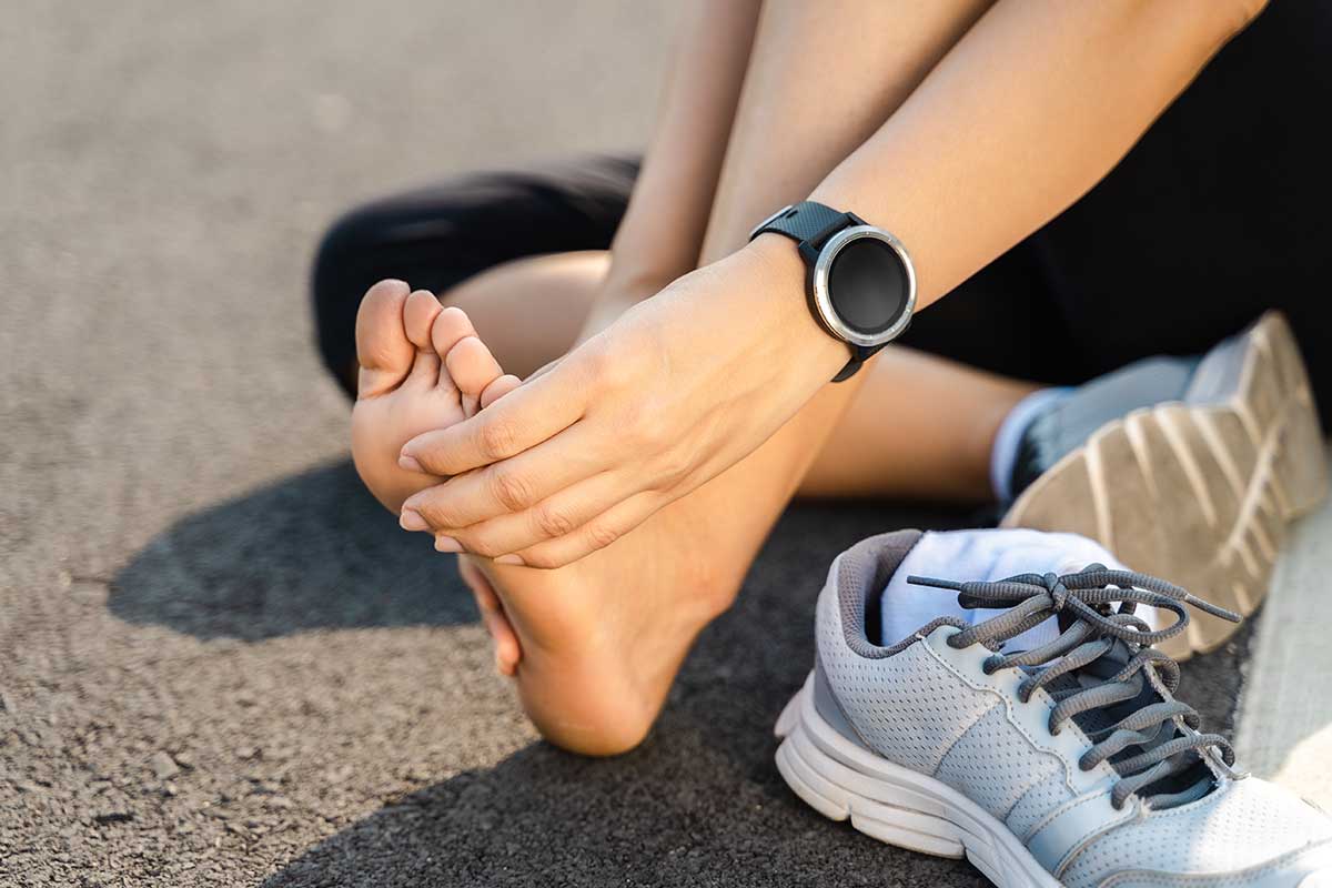 A person sits on the pavement, massaging their bare foot while wearing a smartwatch—a testament to technology meeting fitness. Gray athletic shoes and white socks lie nearby, suggesting a break from exercise, likely prompted by one of the 5 common causes of foot pain experienced by runners.