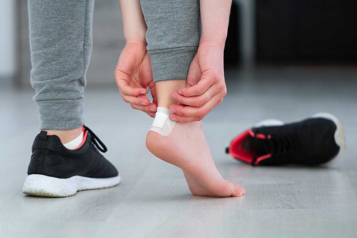 A person is applying a small white bandage to the back of their foot near the ankle, likely addressing one of the common foot and ankle conditions seen in Central Arkansas. They are wearing gray leggings, with one black athletic shoe with a white sole lying on the light-colored wood floor nearby.