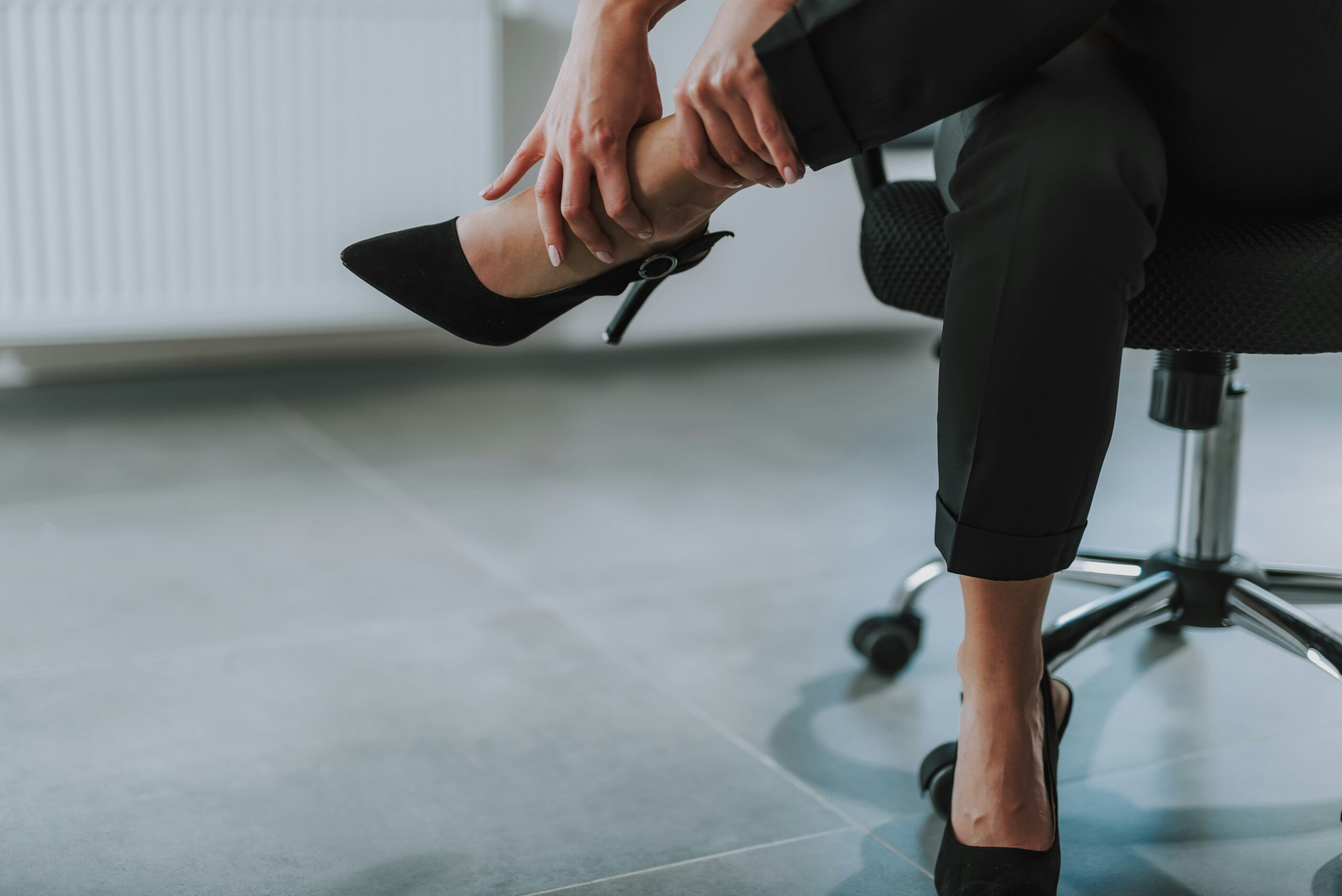 A person sitting on an office chair holds their ankle in discomfort while wearing black high heels and black pants, suggesting foot pain or injury.