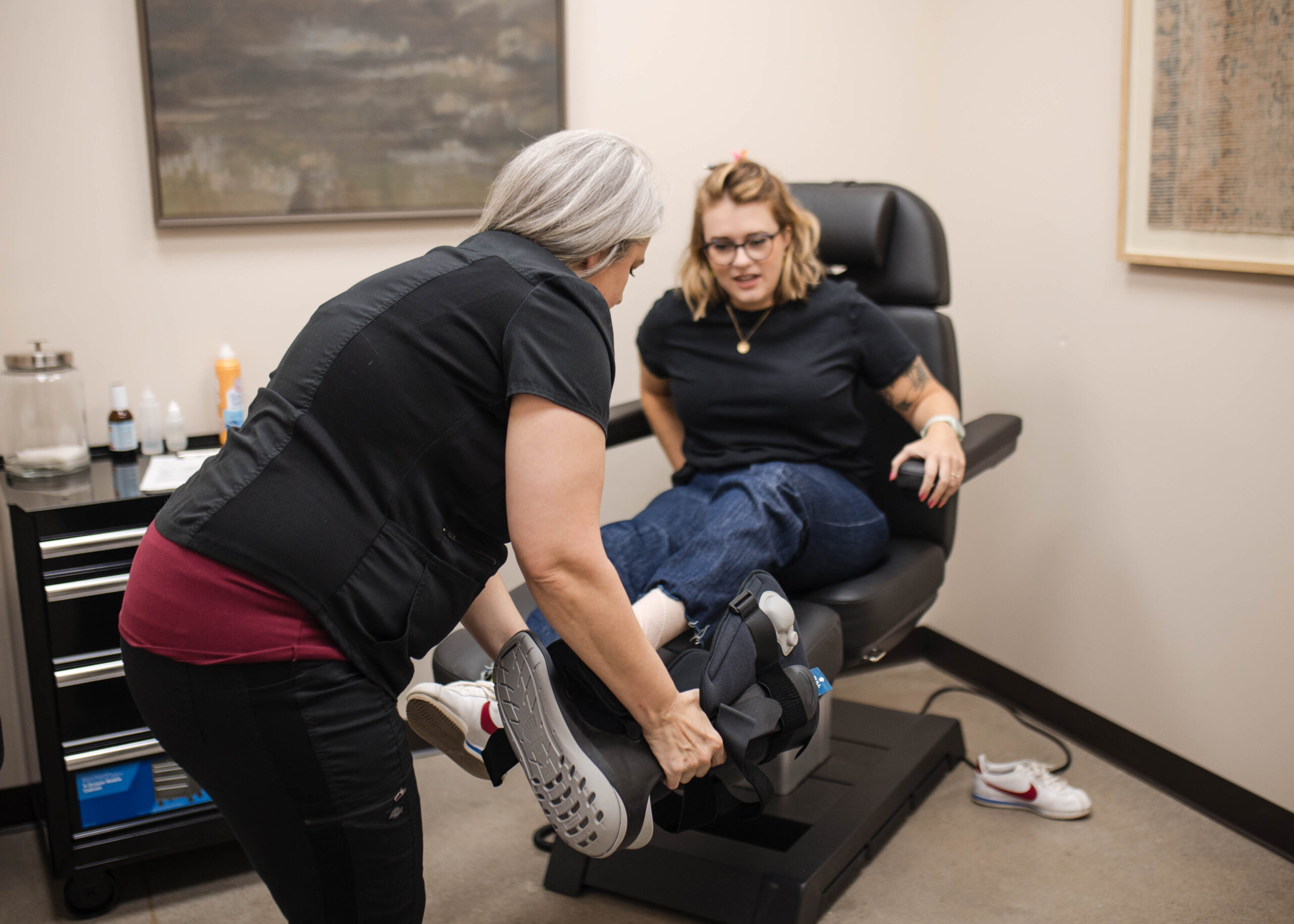 A healthcare professional helps a woman adjust her orthopedic walking boot while she sits on a medical exam chair in a clinic room.