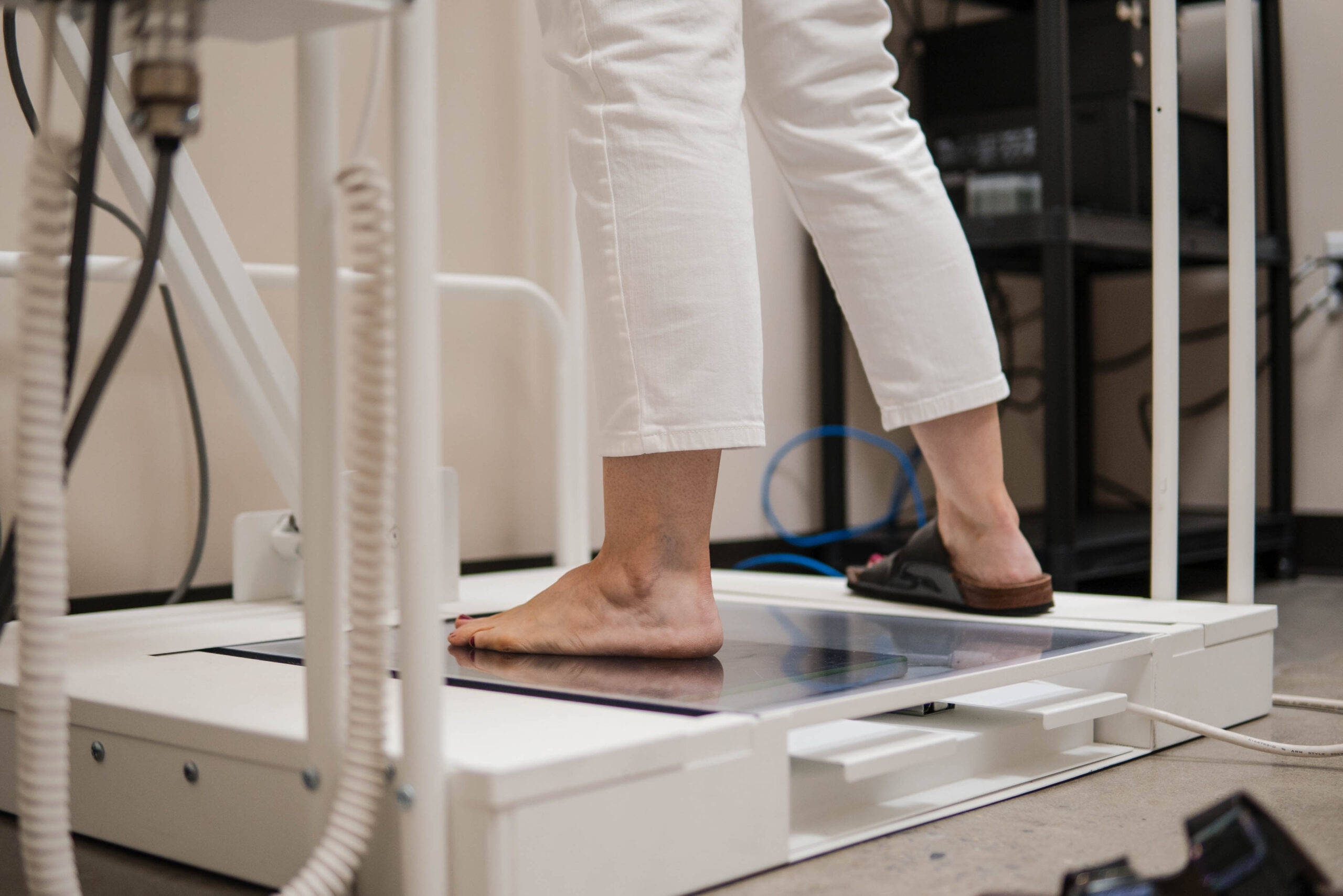A person wearing white pants stands barefoot on a medical imaging device, possibly for a foot or leg scan, in a clinical setting.