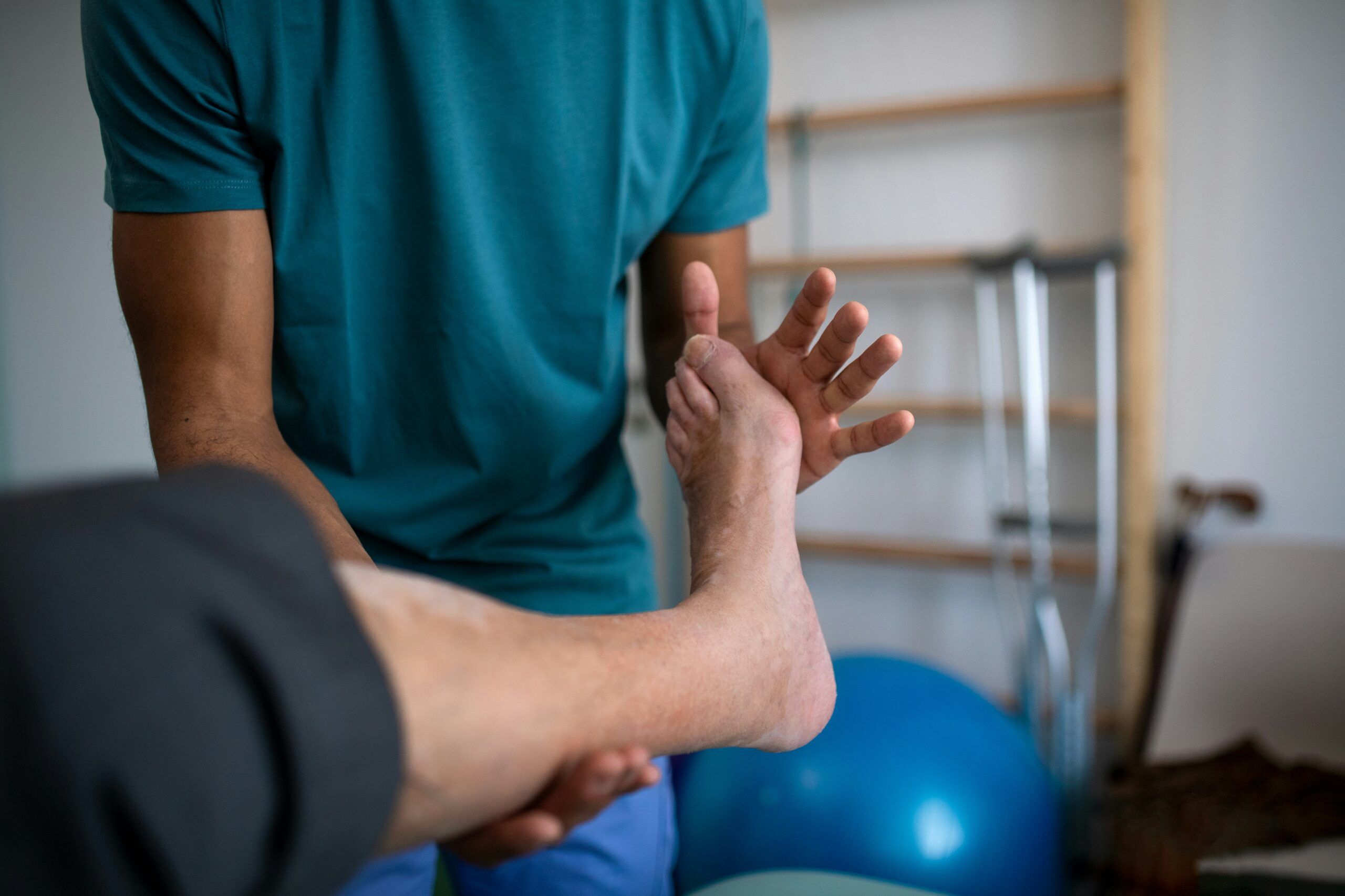 A person in a teal shirt supports and gently moves another person’s foot during a physical therapy session. Exercise equipment and crutches are visible in the background.