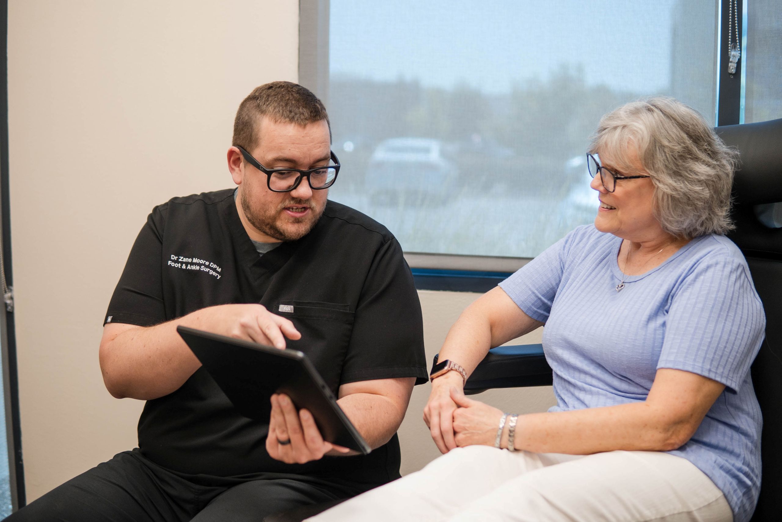 A male healthcare professional in black scrubs shows information on a tablet to an older woman with high arches, who is smiling while sitting in a clinic chair near a window.