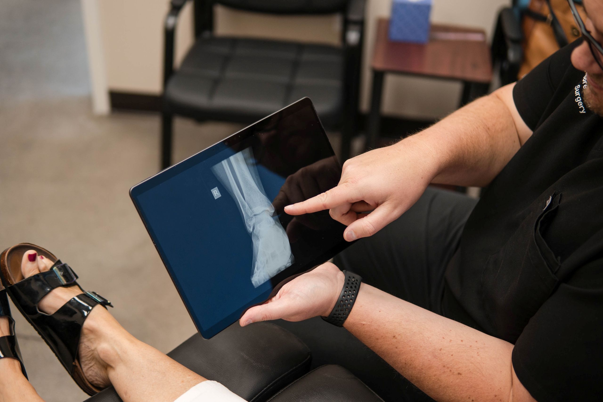 A person points to an X-ray image of an ankle displayed on a tablet, while another person’s foot with black sandals—possibly affected by bunions—rests nearby, suggesting a medical consultation.