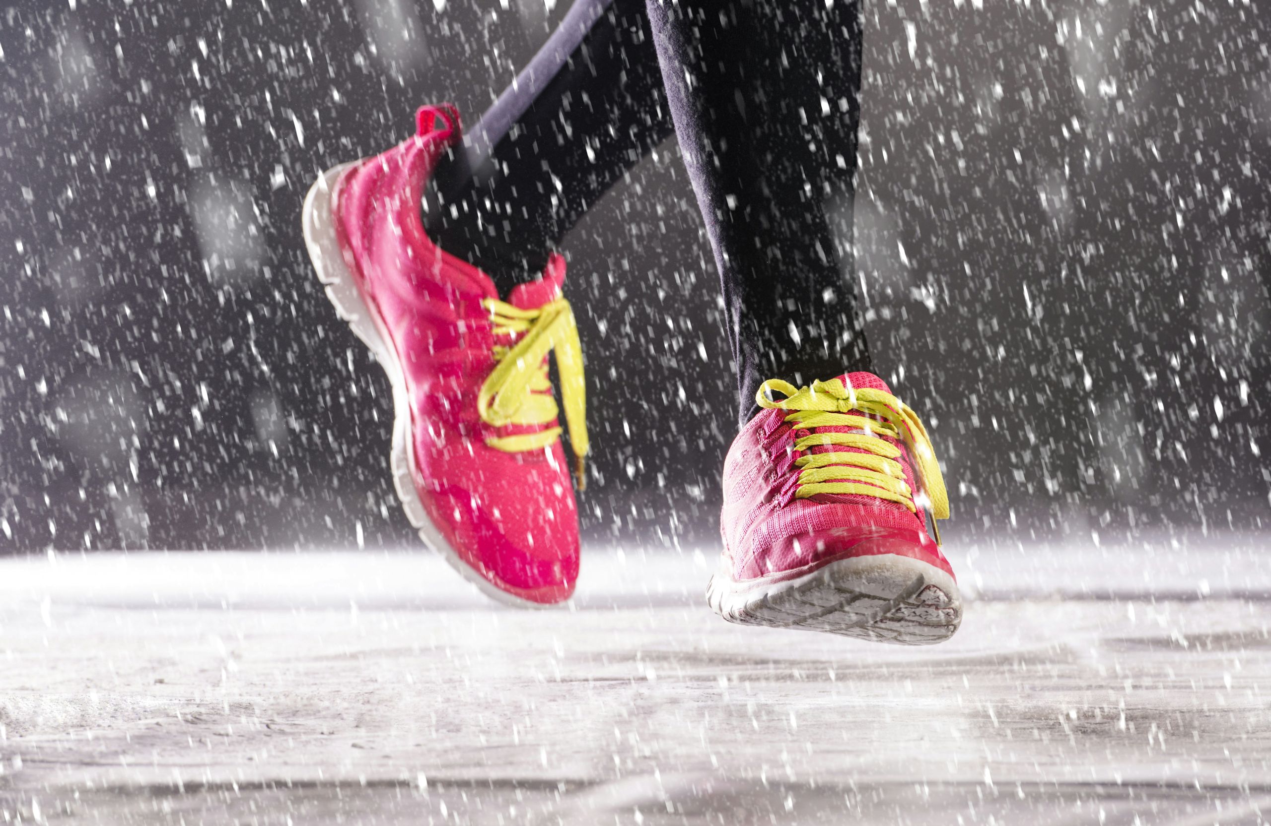 Close-up of a person wearing bright pink running shoes with yellow laces, running outdoors in the rain. Water splashes around their feet as they move along a wet surface—avoid cold weather foot pains with proper footwear.