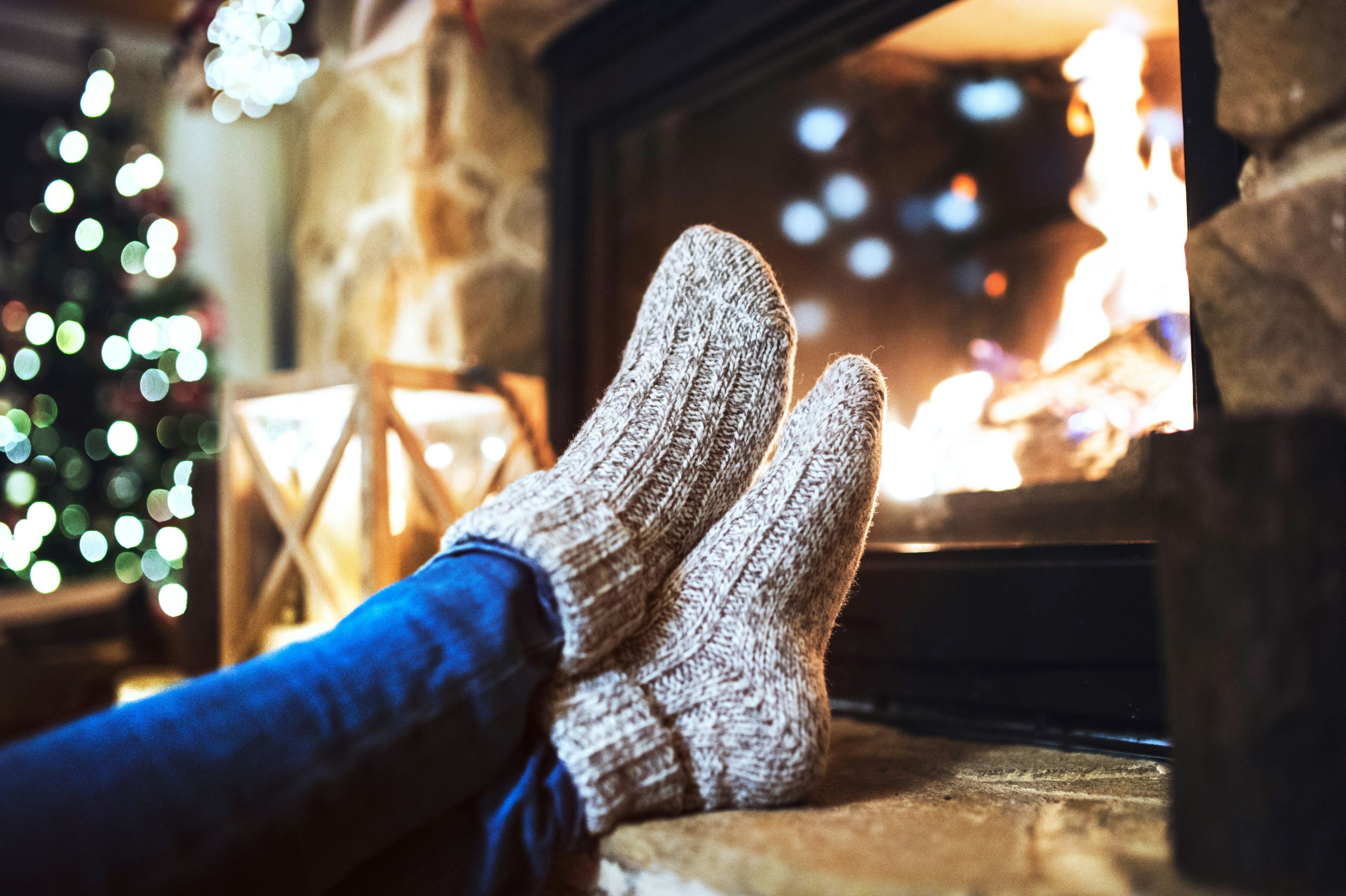 A person with poor circulation and cold feet wears cozy knitted socks, resting near a fireplace with a glowing fire and a blurred Christmas tree in the background, creating a warm, festive atmosphere.