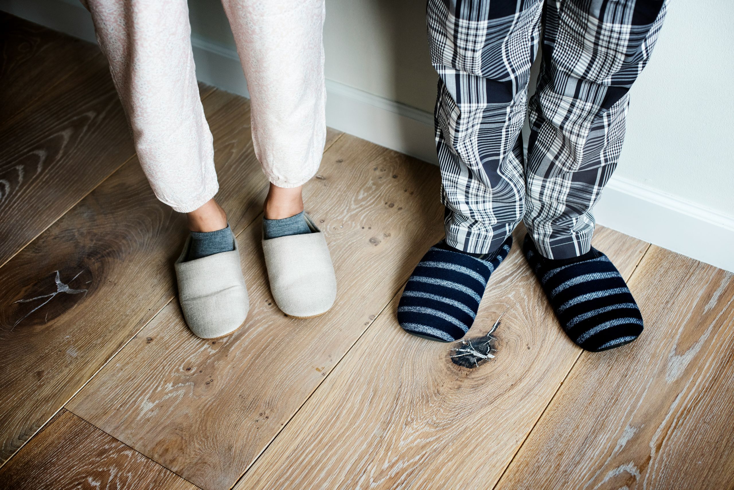 Two people stand side by side on a wooden floor, wearing pajamas and slippers that offer comfort and support for foot health—one in light pajamas with beige slippers, the other in plaid pajamas with dark striped slippers.
