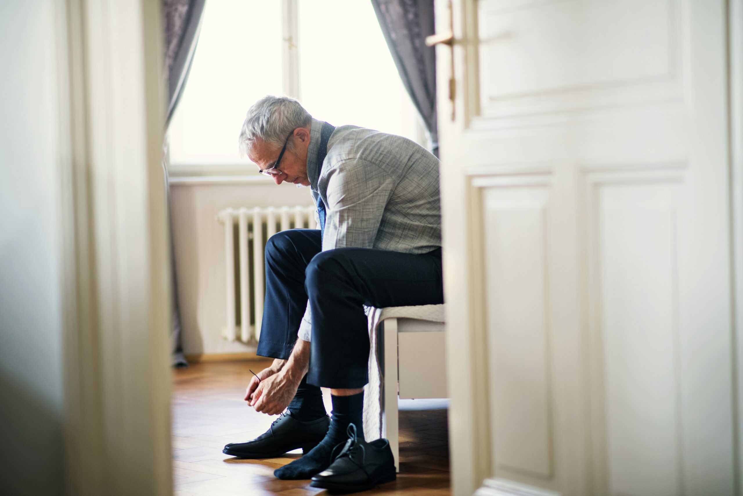 An older man sits on a bench in a bright room, bending forward with care to put on black dress shoes, perhaps feeling some morning foot pain. He wears a gray jacket, dark pants, and glasses as natural light streams through the window behind him.