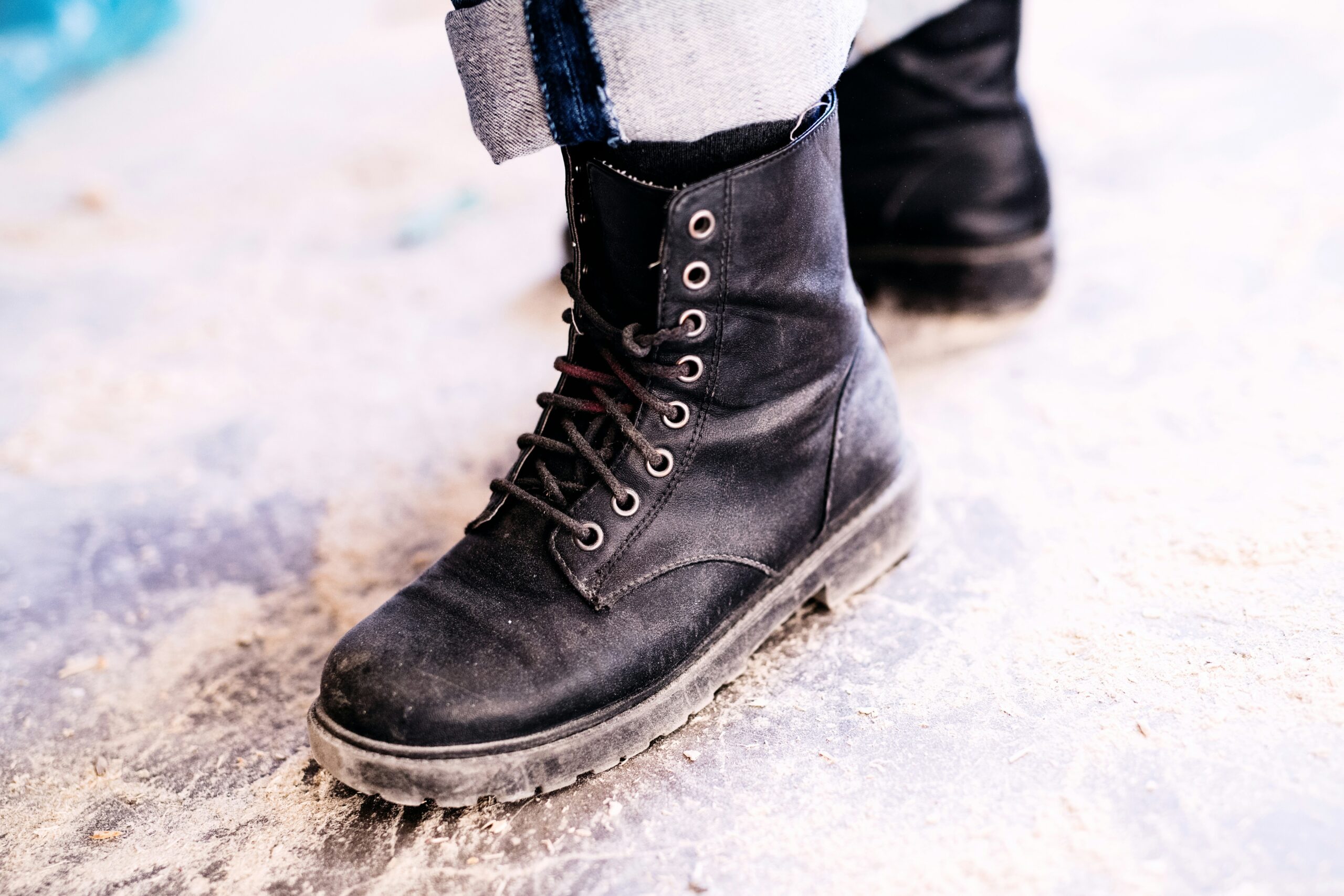 A close-up of a person wearing black lace-up boots and light-colored rolled-up jeans, standing on a dusty, textured floor.