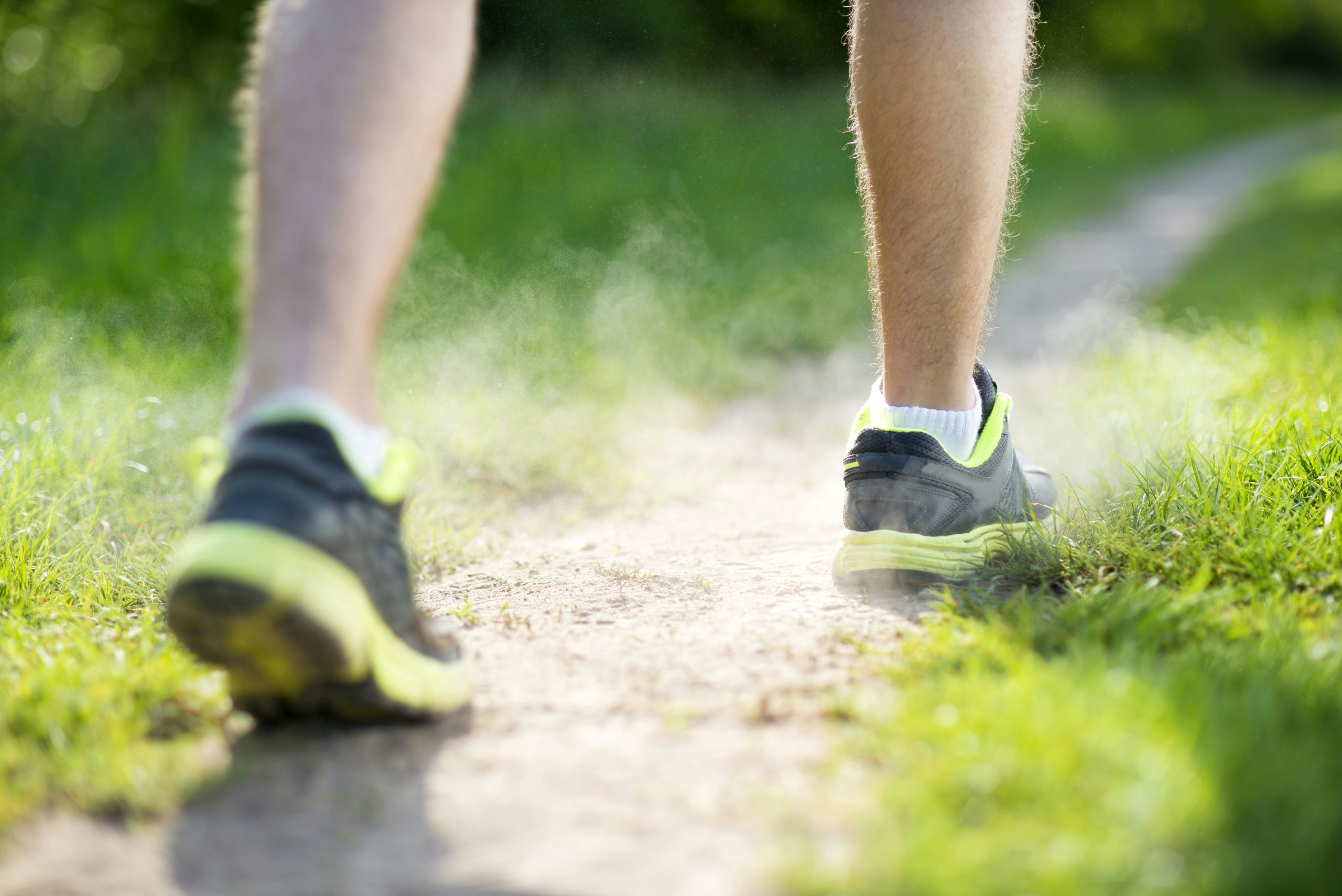 Close-up of a persons legs as they run on a dirt path, kicking up dust. The focus is on their athletic shoes and the surrounding green grass.