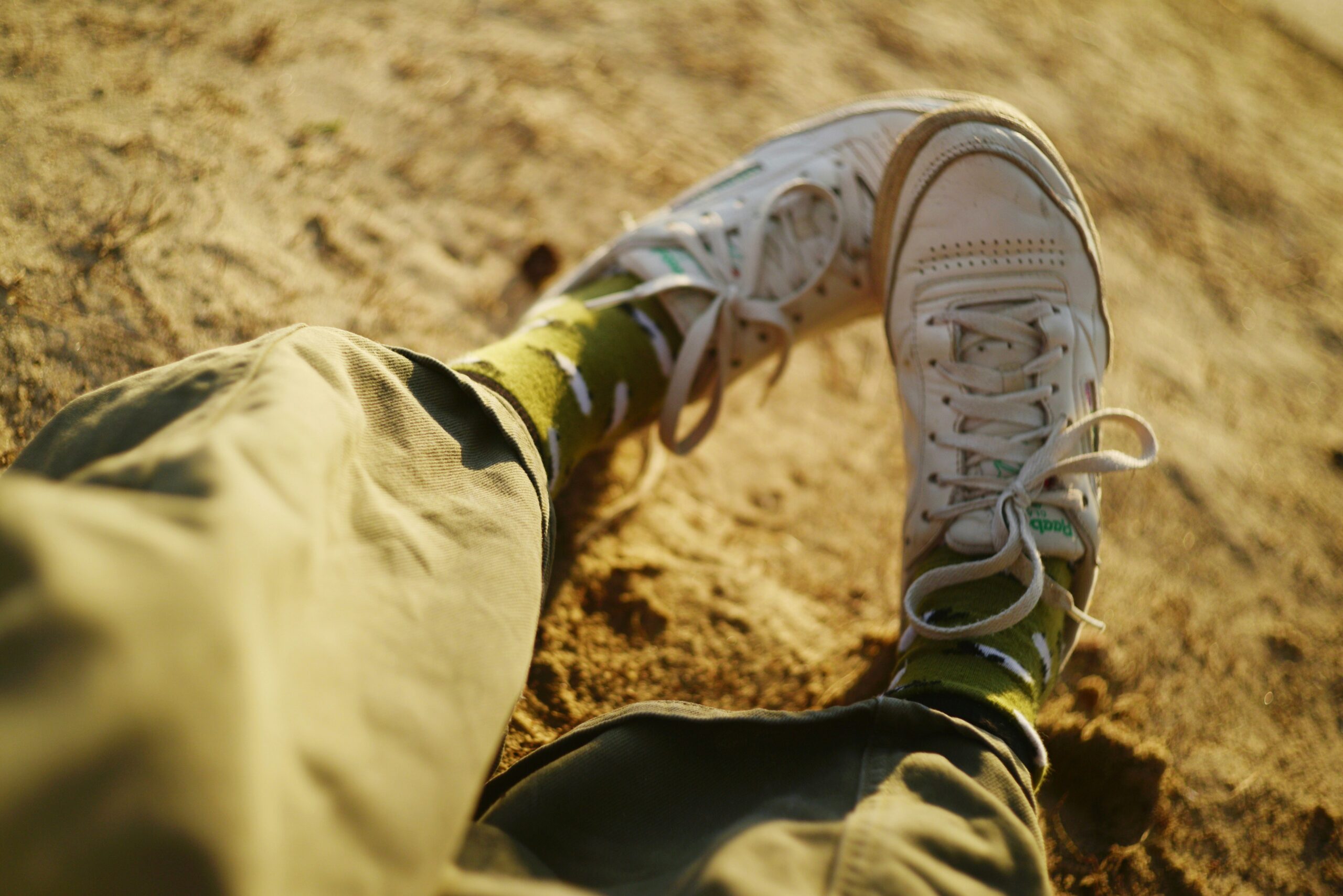 foot care tips (2) Person wearing green cactus-patterned socks and white sneakers, sitting on sandy ground with legs crossed. The image is taken from their perspective, showing their lower legs and feet—perfect for sharing foot care tips in the warm, soft sunlight.