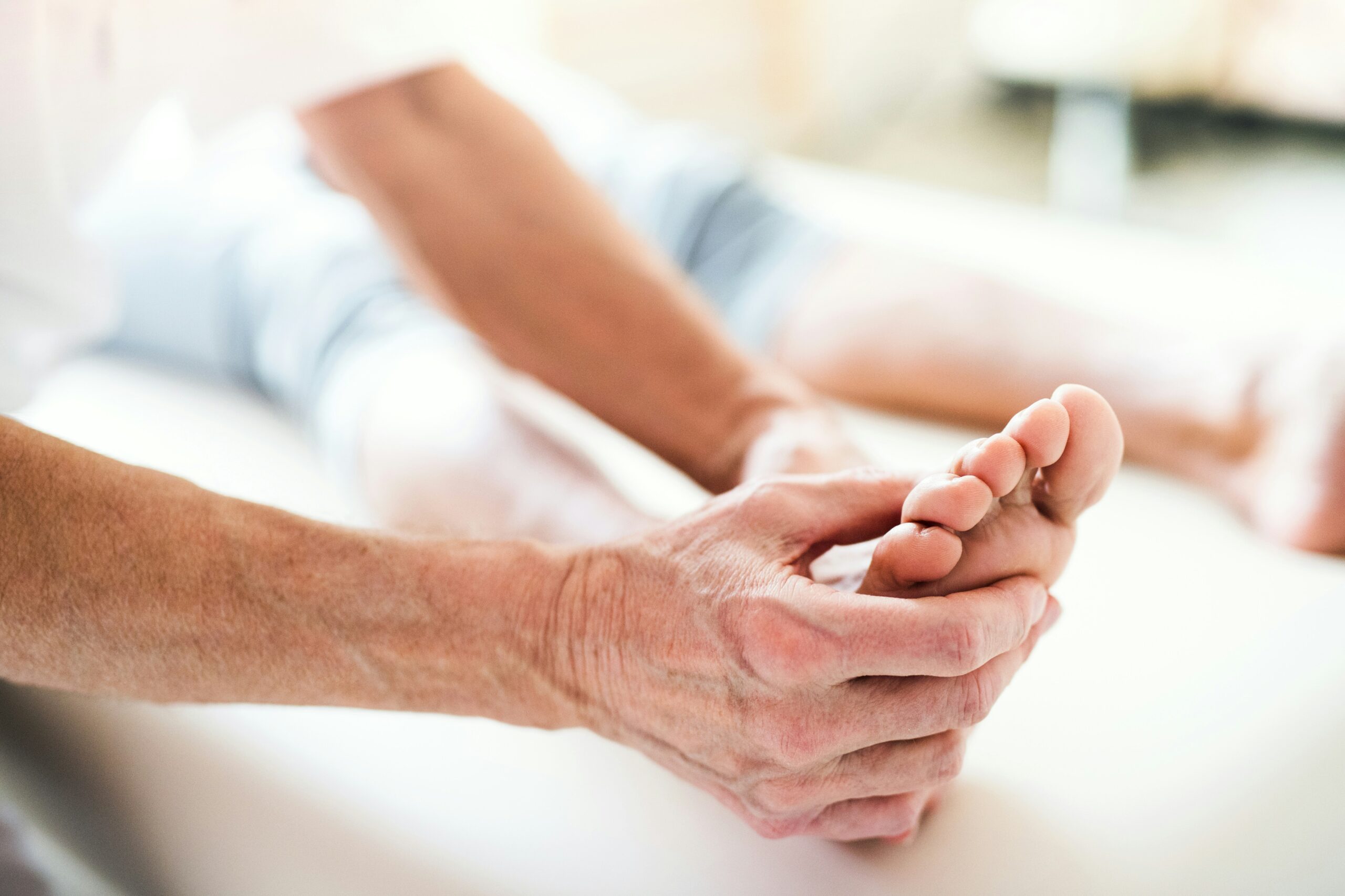 Close-up of a person sitting with legs extended, using both hands to gently stretch and massage the toes of one foot, suggesting improved foot circulation and relaxation as part of a soothing foot care routine.
