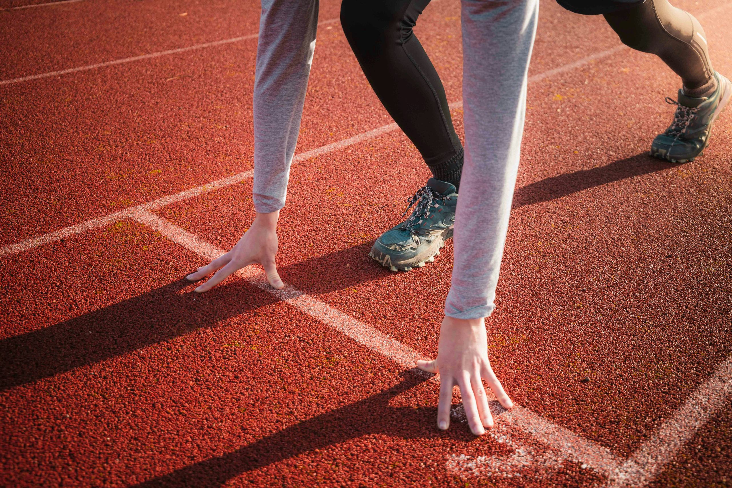 Close-up of a runner in a starting position on a red track, hands on the ground and one knee bent, ready to sprint. The persons face is not visible.