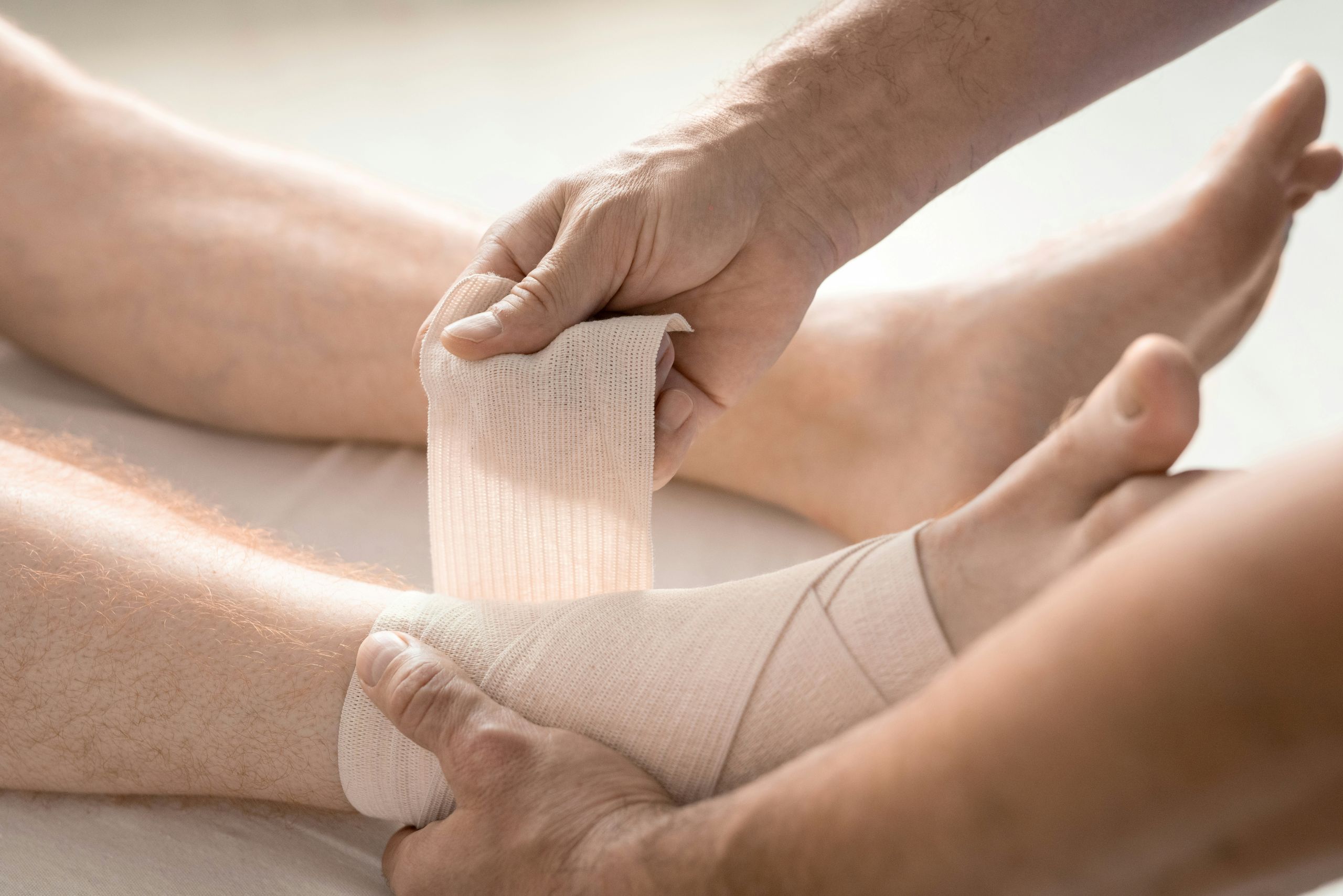 Why Your Ankles Keep Rolling and How to Improve Stability A close-up of hands wrapping a bandage around a persons injured ankle, providing support and care. The person is lying down with their leg extended.