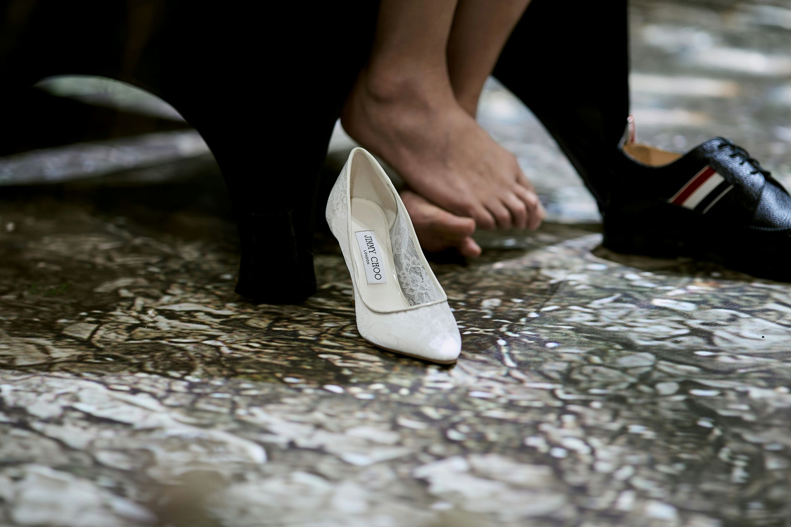 A person sits barefoot next to a white high heel labeled Jimmy Choo and a black sneaker with red and white stripes, all on a marbled floor.