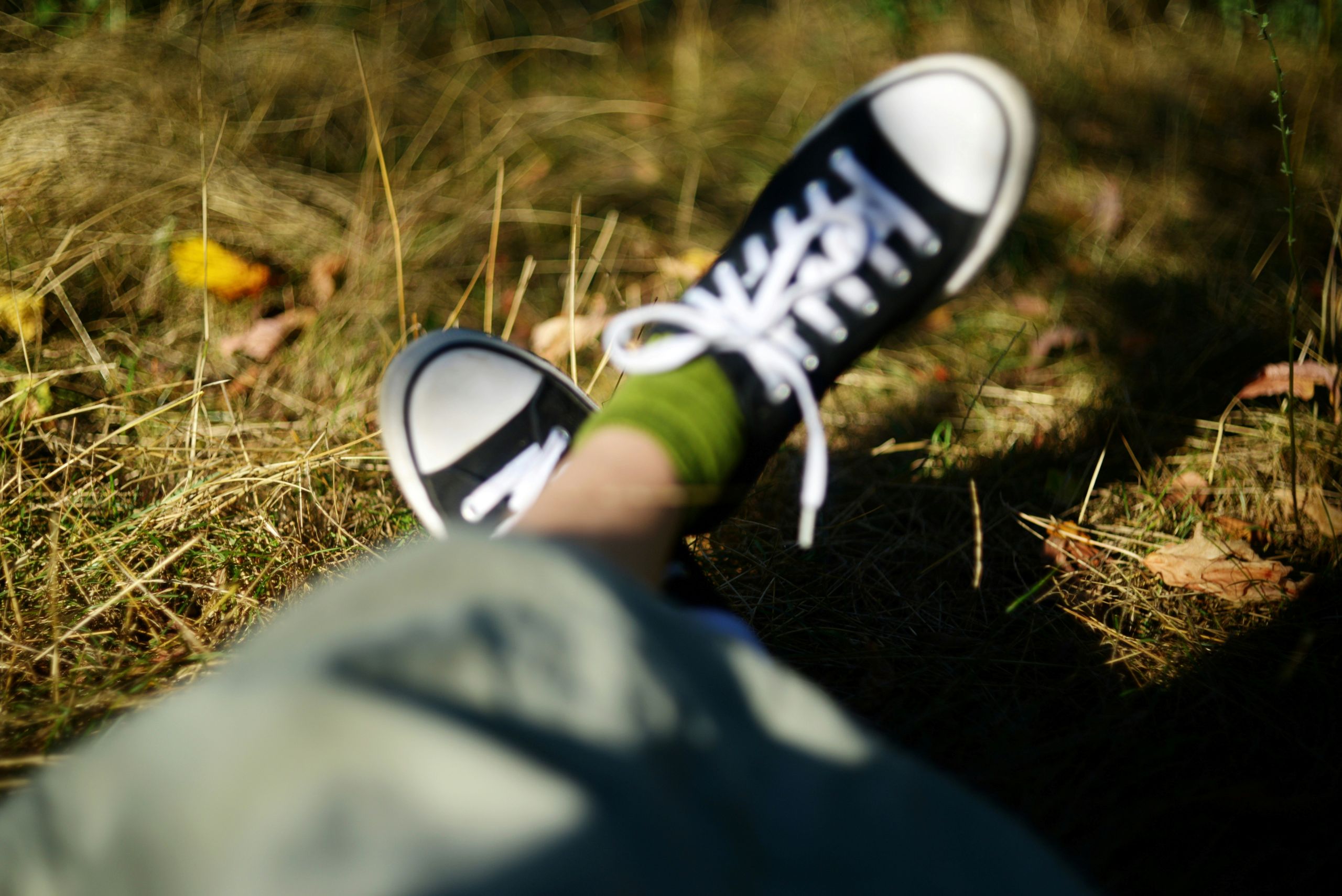A person wearing green socks and black-and-white sneakers sits on dry grass, with the photo focusing on their shoes and blurred background.