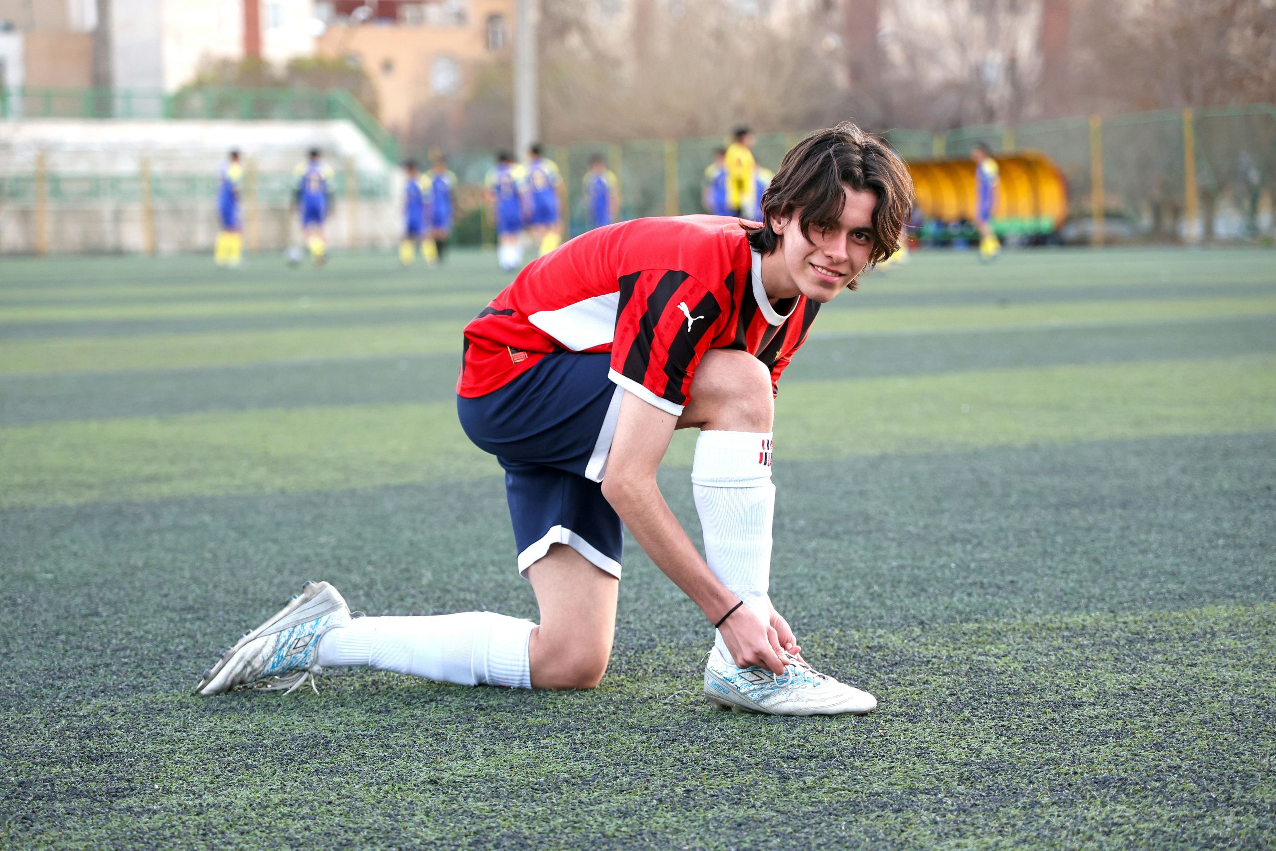 A smiling soccer player kneels on a field while tying their cleats.